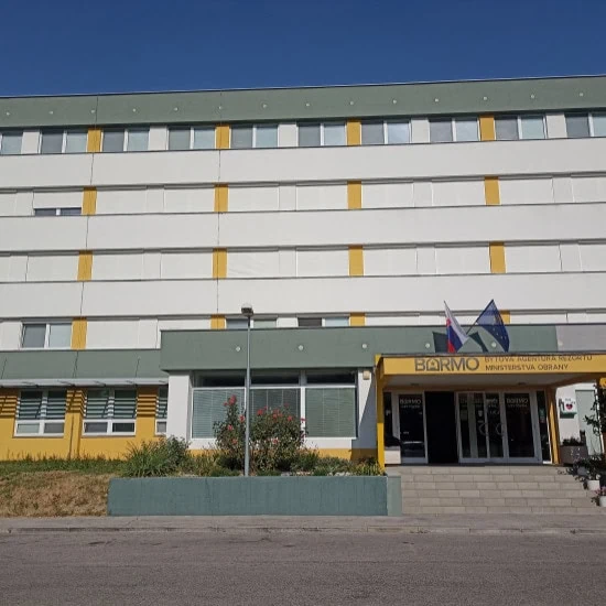 Štvorpodlažná budova s bielo-žltou fasádou, s hlavným vchodom s baldachýnom. Nad vchodom stoja dva stožiare s vlajkami a nad dverami je viditeľný nápis BÁRMO. Vpredu sú kríky a pouličná lampa.